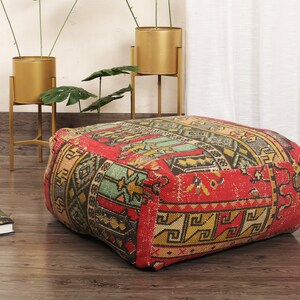 May include: A red, green, and yellow patterned square ottoman with a fringe border. The ottoman is made of fabric and has a traditional design.