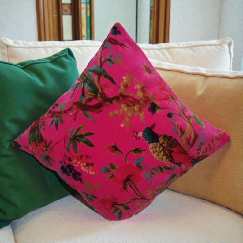 May include: A hot pink square pillow with a bird and floral pattern. The pillow has a velvet texture and is surrounded by other pillows in green and tan.