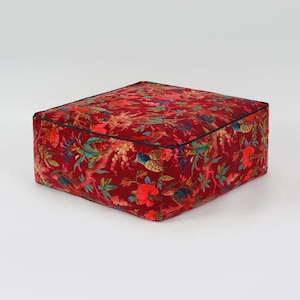 May include: A red velvet square ottoman with a floral and bird print. The ottoman has a black trim around the edges.