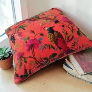 Pode incluir: Almofada decorativa quadrada com um fundo vermelho vibrante, apresentando um padrão floral e de pássaros detalhado em tons de rosa, verde e azul. A almofada está colocada perto de uma janela, com uma pequena suculenta em vaso e uma pilha de livros.