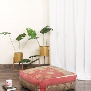 May include: A square, patterned floor cushion with a red and cream floral design. The cushion is sitting on a wooden floor in front of a white wall and a white curtain.