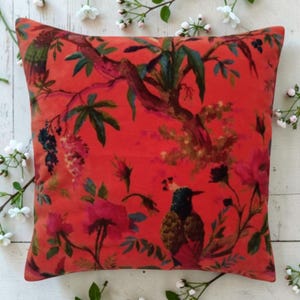 May include: Square decorative pillow with a vibrant red background featuring a botanical design. The pattern includes birds, flowers, and foliage in shades of pink, green, and blue. The pillow is a square shape, perfect for adding a pop of color to any room.