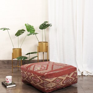 May include: A red and beige patterned floor cushion with a geometric design. The cushion is sitting on a wooden floor in front of a white curtain and potted plants.