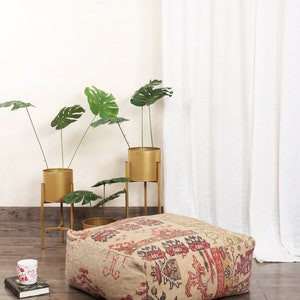 May include: A beige, square, patterned floor cushion with red, black, and white accents. The cushion is sitting on a wooden floor in front of a white wall and a white curtain.