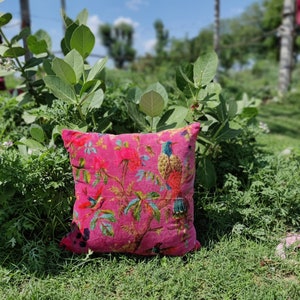 May include: A pink throw pillow with a floral and bird print. The pillow is sitting on a bed of green grass.