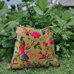 May include: A square throw pillow with a floral and bird print. The pillow is a light brown color with pink flowers, green leaves, and colorful birds. The pillow is sitting on a bed of green grass.