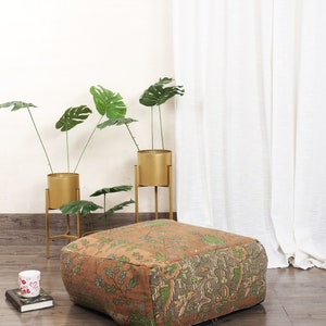 May include: A square, brown and green patterned floor cushion with a zipper closure. The cushion is sitting on a wooden floor in front of a white curtain and two gold plant stands with green plants.