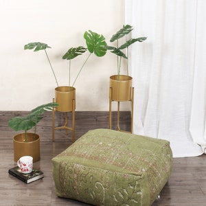 May include: A green square floor cushion with a patterned design. Three gold metal plant stands with green leafy plants in gold pots.