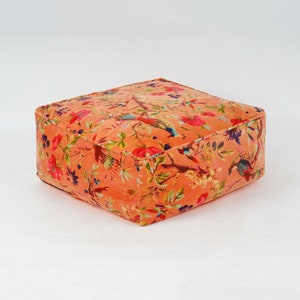 May include: A square, peach-colored ottoman with a floral print featuring birds and flowers. The ottoman is made of a soft, plush fabric.