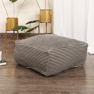 May include: A square floor cushion featuring a woven fabric with a pattern of thin, alternating black and off-white horizontal stripes. The cushion has a structured, boxy shape with defined edges and a textured, natural fiber appearance throughout.