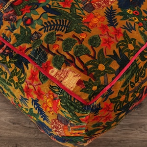 May include: A square, mustard yellow velvet cushion with a colorful floral and botanical print. The cushion has a red trim around the edges.