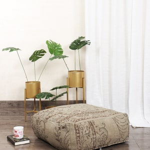 May include: A large, square, beige ottoman with a brown patterned design. The ottoman is sitting on a wooden floor in front of a white curtain. There are three gold metal plant stands with green plants in them.