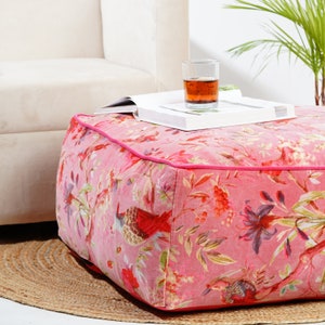 May include: A pink floral patterned ottoman with a pink trim. The ottoman is sitting on a woven rug. A glass of iced tea and a book are on top of the ottoman.