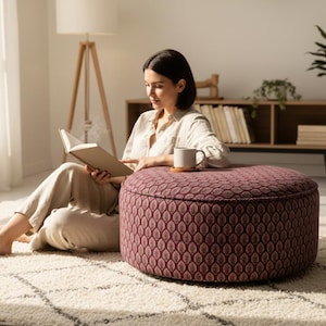 Puede incluir: Una mujer lee un libro sentada en una alfombra estampada junto a una otomana burdeos. Una taza descansa sobre un posavasos de madera. La habitación tiene una paleta de colores neutros con una estantería de madera y una lámpara de pie.