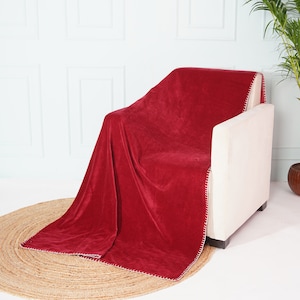 Puede incluir: Una manta de terciopelo rojo con costuras blancas drapeada sobre un sillón blanco. La manta es suave y mullida, y las costuras añaden un toque decorativo.