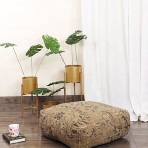 May include: A square, brown and black patterned floor cushion with a floral design. The cushion is sitting on a wooden floor in front of a white wall and a white curtain. There are three gold metal plant stands with potted plants in the background.