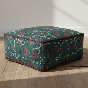 May include: Square ottoman with a floral pattern in shades of teal, red, and cream. The edges are trimmed with a dark red piping. The ottoman is a square shape and sits on a light brown surface.