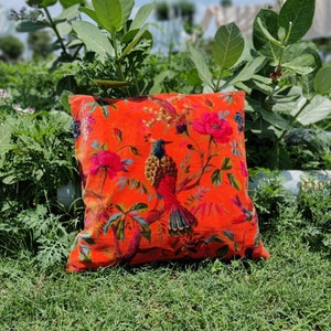 Puede incluir: Almohada de terciopelo naranja con estampado floral y de pájaros. La almohada presenta un pájaro grande con una cola larga y un pájaro más pequeño en vuelo. Las flores son rosas y blancas.