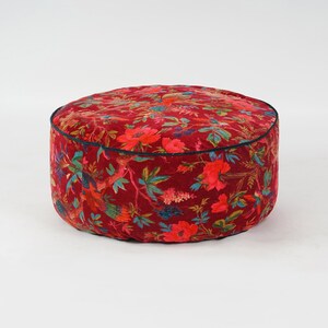 May include: A round red velvet ottoman with a floral and bird print. The ottoman has a black trim around the top edge.