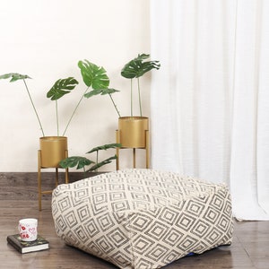 May include: A white and black geometric patterned floor cushion with a square shape. The cushion is sitting on a wooden floor in front of a white wall and a white curtain.