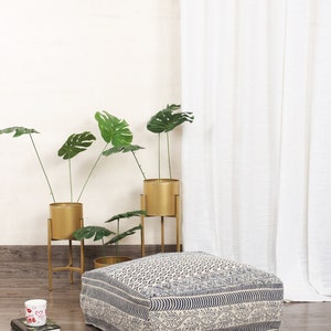 May include: A blue and white patterned floor cushion with a geometric design. The cushion is sitting on a wooden floor in front of a white wall and a white curtain. There are three gold metal plant stands with green plants in gold pots.