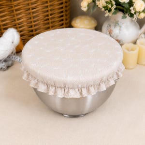 May include: A round, beige fabric bowl cover featuring a subtle white leaf pattern and a ruffled edge. The cover is fitted securely over the top of a stainless steel bowl, providing a snug fit around the rim with its elasticated construction.