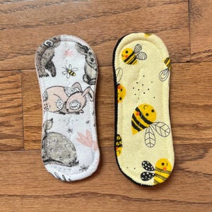 May include: Two reusable cloth pads. One features a white background with bunny and bee illustrations. The other has a yellow background with bee graphics and a black border. These pads are designed for feminine hygiene.