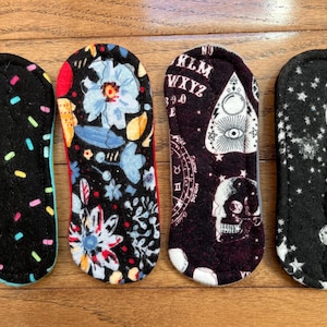 May include: Four reusable cloth menstrual pads with different patterns. One has colorful sprinkles, another has blue floral designs, a third features a skull and Ouija board, and the last has a moon and stars.
