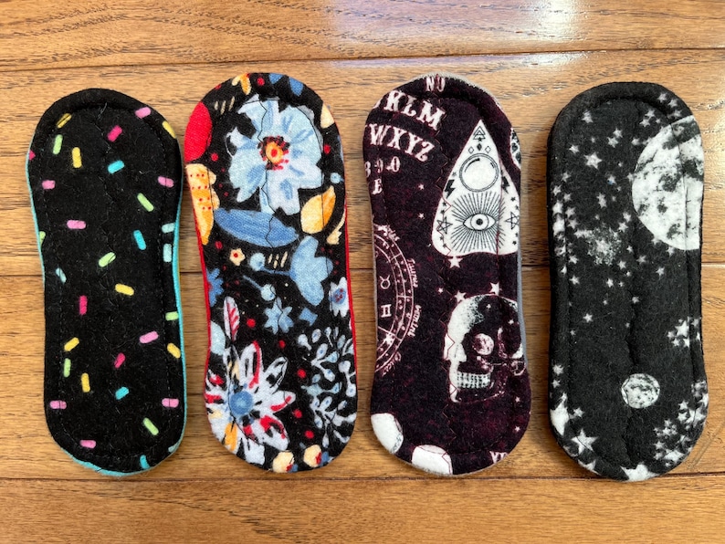 May include: Four reusable cloth menstrual pads with different patterns. One has colorful sprinkles, another has blue floral designs, a third features a skull and Ouija board, and the last has a moon and stars.
