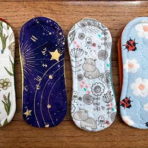 May include: Four reusable cloth menstrual pads with various patterns. Designs include a white floral and mouse print, a navy blue celestial print, a light blue hippo print, and a light blue ladybug print. The pads are rectangular with rounded ends.