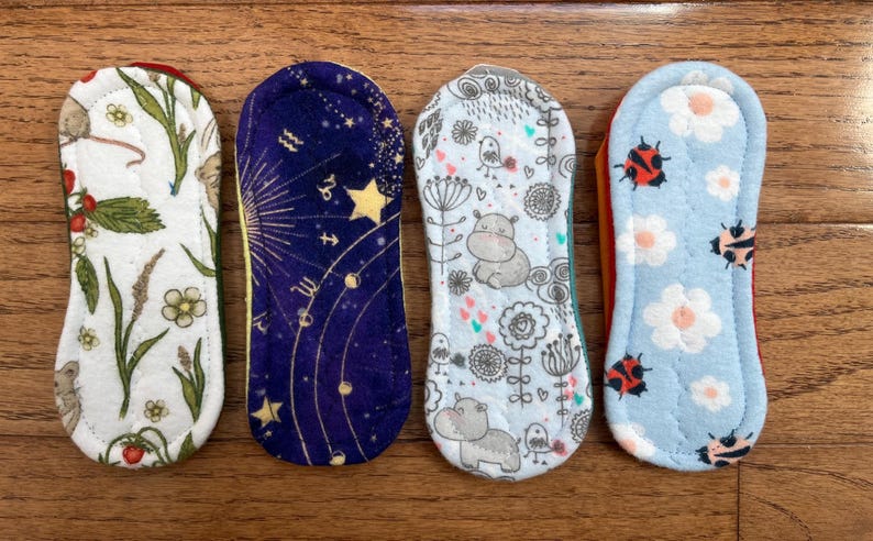 May include: Four reusable cloth menstrual pads with various patterns. Designs include a white floral and mouse print, a navy blue celestial print, a light blue hippo print, and a light blue ladybug print. The pads are rectangular with rounded ends.