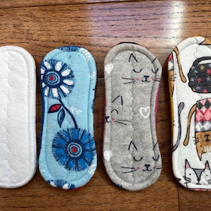 May include: Four reusable cloth menstrual pads. One is white, the second is light blue with a floral design, the third is gray with cat faces, and the fourth is white with cats and geometric patterns.