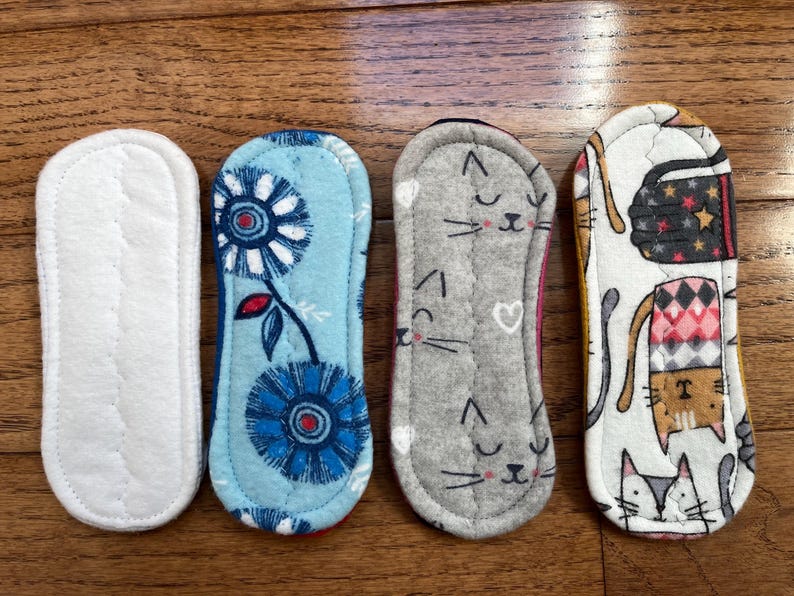 May include: Four reusable cloth menstrual pads. One is white, the second is light blue with a floral design, the third is gray with cat faces, and the fourth is white with cats and geometric patterns.