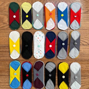 May include: A collection of reusable cloth menstrual pads in various colors and patterns. Each pad features a unique design with contrasting fabric accents and button closures. The pads are arranged on a wooden surface.