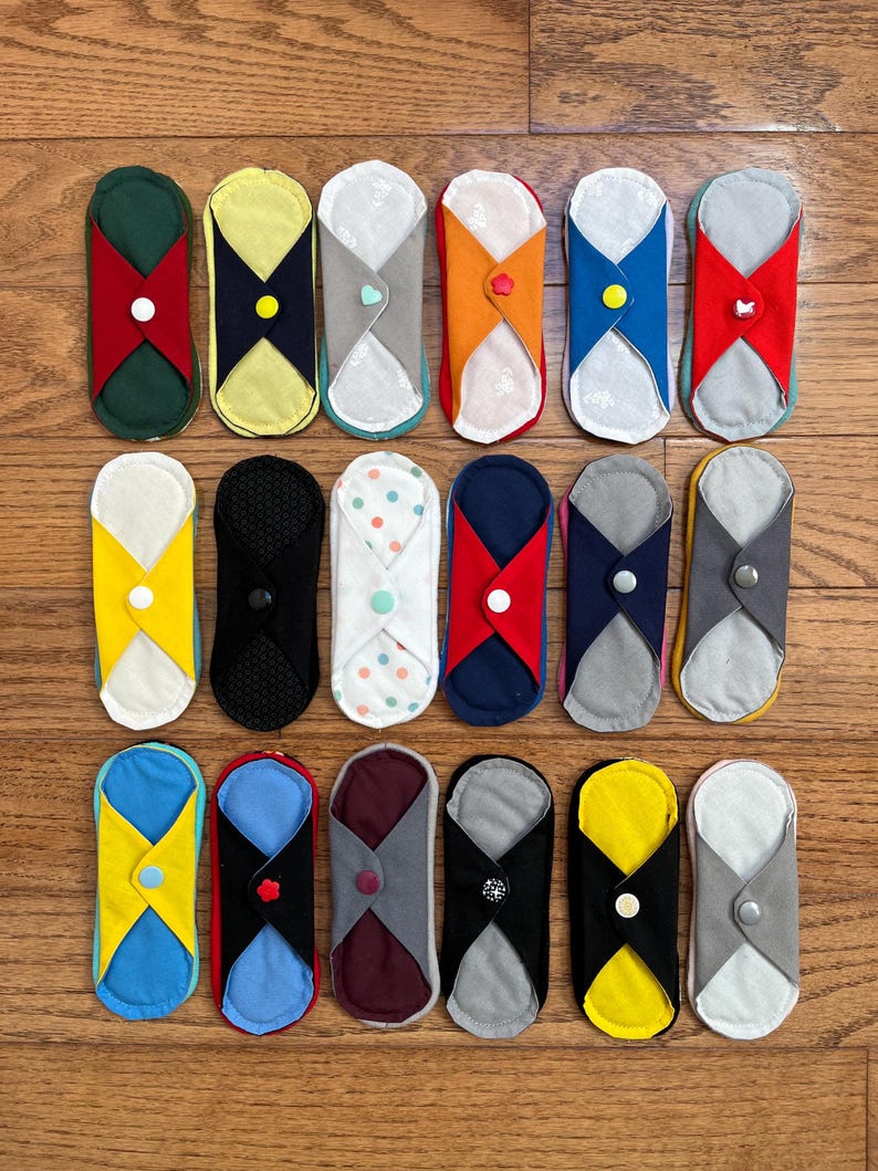 May include: A collection of reusable cloth menstrual pads in various colors and patterns. Each pad features a unique design with contrasting fabric accents and button closures. The pads are arranged on a wooden surface.