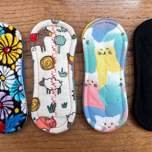 May include: Four reusable cloth menstrual pads. Designs include a floral pattern, farm animals, cats, and a solid black pad. The pads are rectangular with rounded ends and are made of soft fabric.