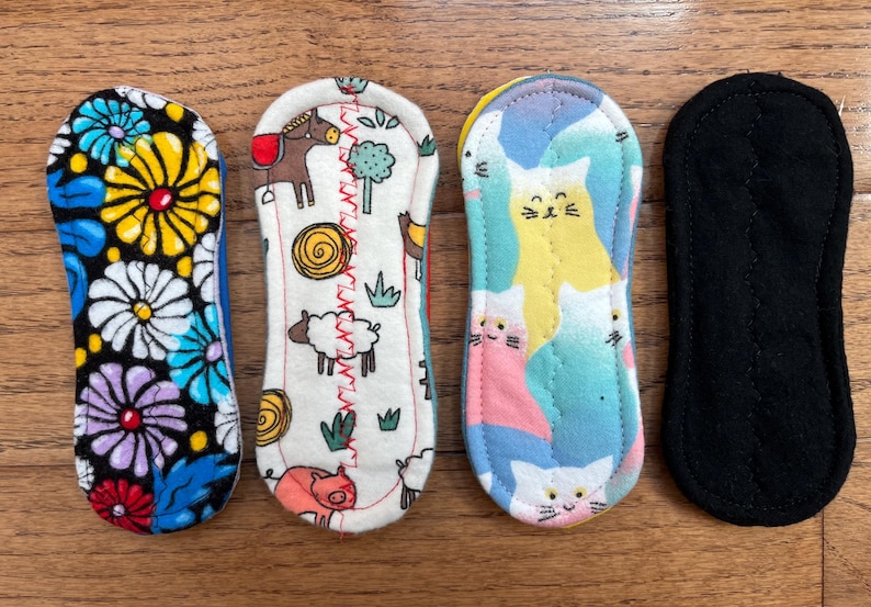 May include: Four reusable cloth menstrual pads. Designs include a floral pattern, farm animals, cats, and a solid black pad. The pads are rectangular with rounded ends and are made of soft fabric.