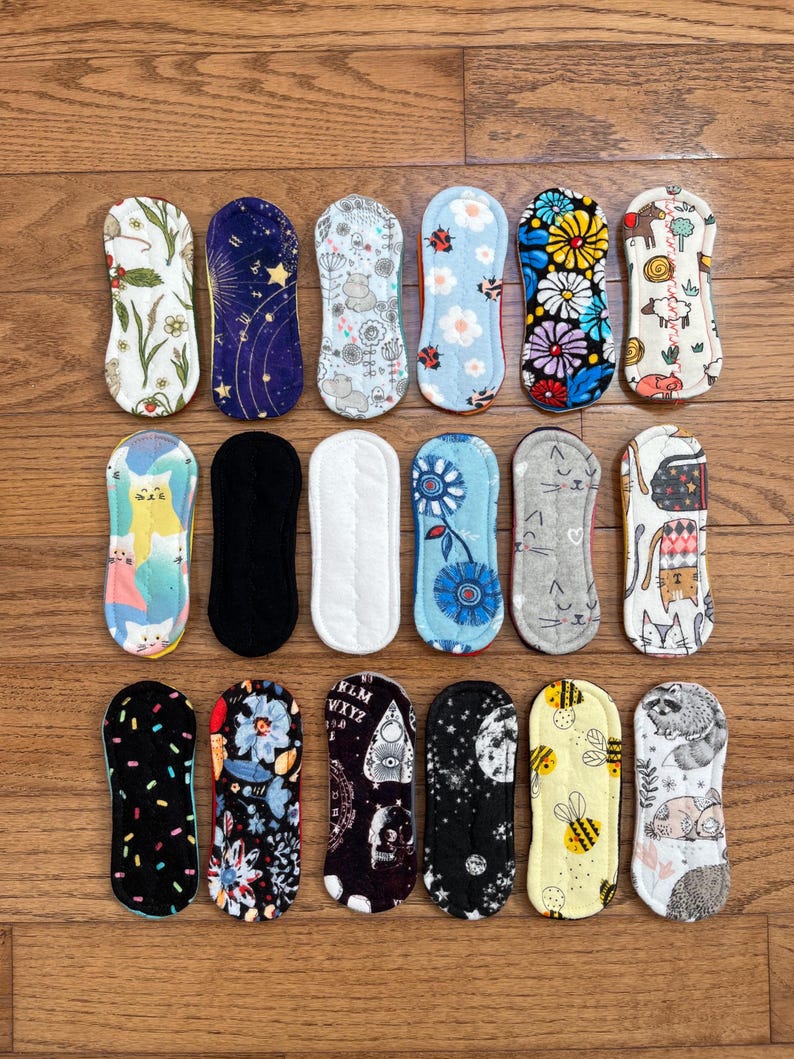 May include: A collection of reusable cloth menstrual pads with various colorful patterns. Designs include floral, celestial, animal, and geometric prints. The pads are arranged on a wooden surface.