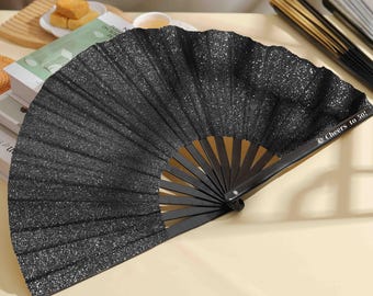 以下が含まれることがあります： キラキラ光る黒い折りたたみ式の扇子で、木製のハンドルが付いています。扇子のハンドルには「Cheers to 50!」という文字が刻まれています。
