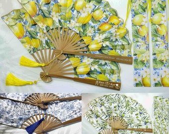 May include: Set of three hand fans with floral designs. The fans are made of bamboo and fabric. The fabric is printed with lemons and flowers on a white background. The fans have yellow tassels. The fans are packaged in matching fabric bags. The bags are printed with lemons and flowers on a white background. The bags have yellow tassels.