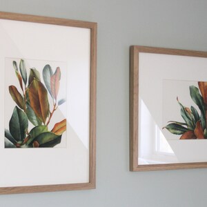 May include: Two framed botanical prints featuring watercolor illustrations of magnolia leaves in shades of green, brown, and red.