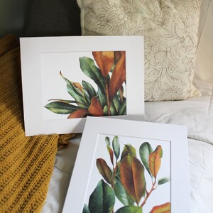 May include: Two framed botanical prints of magnolia leaves in shades of green and brown. The prints are displayed on a white bedspread with a yellow knitted throw blanket.