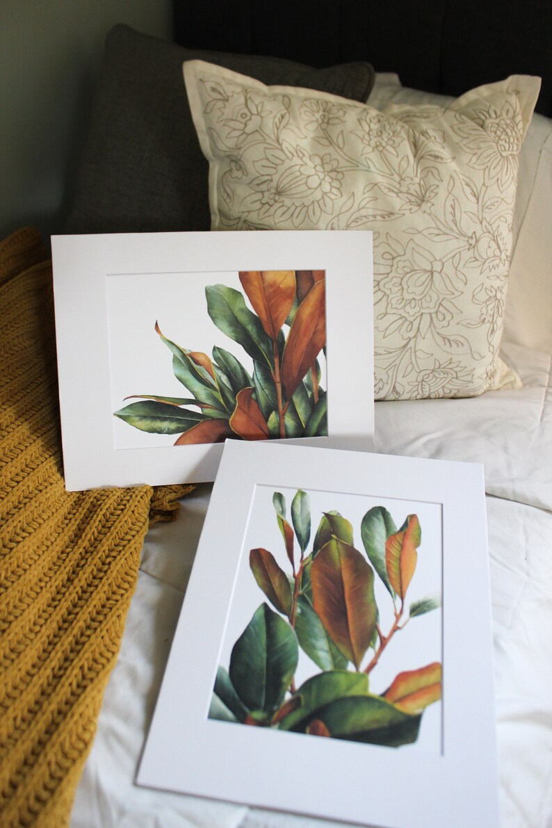 May include: Two framed botanical prints of magnolia leaves in shades of green and brown. The prints are displayed on a white bedspread with a yellow knitted throw blanket.