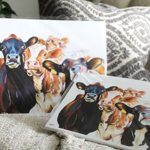 May include: A set of four watercolor paintings of cows. The cows are brown and white with black spots. The paintings are on white paper and are framed in white.