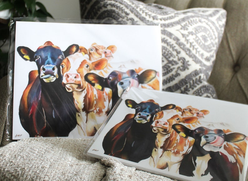 May include: A set of four watercolor paintings of cows. The cows are brown and white with black spots. The paintings are on white paper and are framed in white.