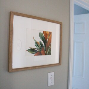 May include: A framed watercolor painting of a leafy plant with green and brown leaves. The painting is in a light wood frame and is hanging on a light gray wall.