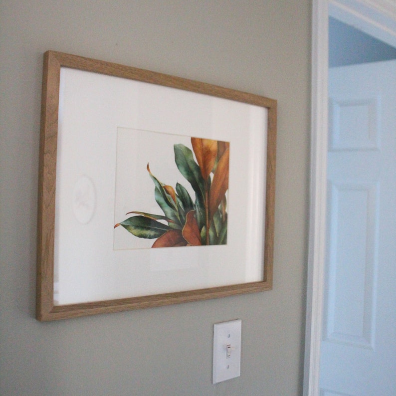 May include: A framed watercolor painting of a leafy plant with green and brown leaves. The painting is in a light wood frame and is hanging on a light gray wall.
