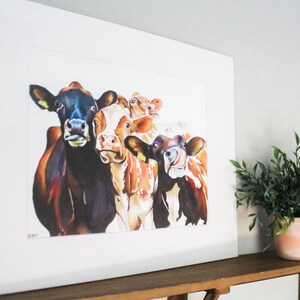 May include: A watercolor painting of five cows with different colored markings. The cows are looking directly at the viewer with their tongues sticking out. The painting is framed in a white frame.