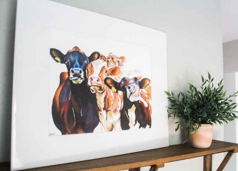 May include: A watercolor painting of five cows with different colored markings. The cows are looking directly at the viewer with their tongues sticking out. The painting is framed in a white frame.