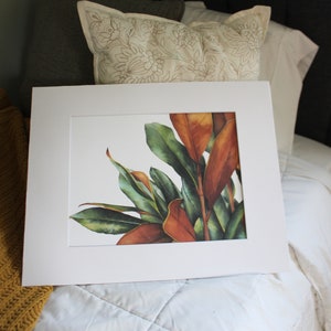 May include: A framed watercolor painting of a leafy plant with green and orange leaves. The painting is on a white background and is framed in a white frame.